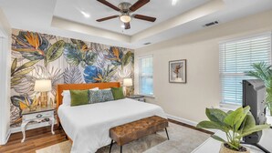 A bedroom with a bed, a ceiling fan, a wall mural, a television, and a potted plant.