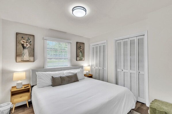 A bedroom with a bed, bedside tables, a window with blinds, and a set of white louvered doors.