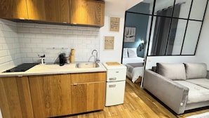 A compact kitchen with wooden cabinets, a white countertop, and a built-in stove. There is a small white refrigerator and a beige sofa in the living area.