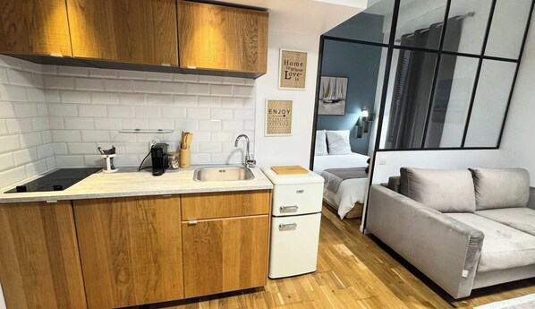 A compact kitchen with wooden cabinets, a white countertop, and a built-in stove. There is a small white refrigerator and a beige sofa in the living area.