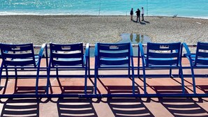 A beach with a row of blue chairs facing the ocean, a few people standing by the water, and a clear sky.