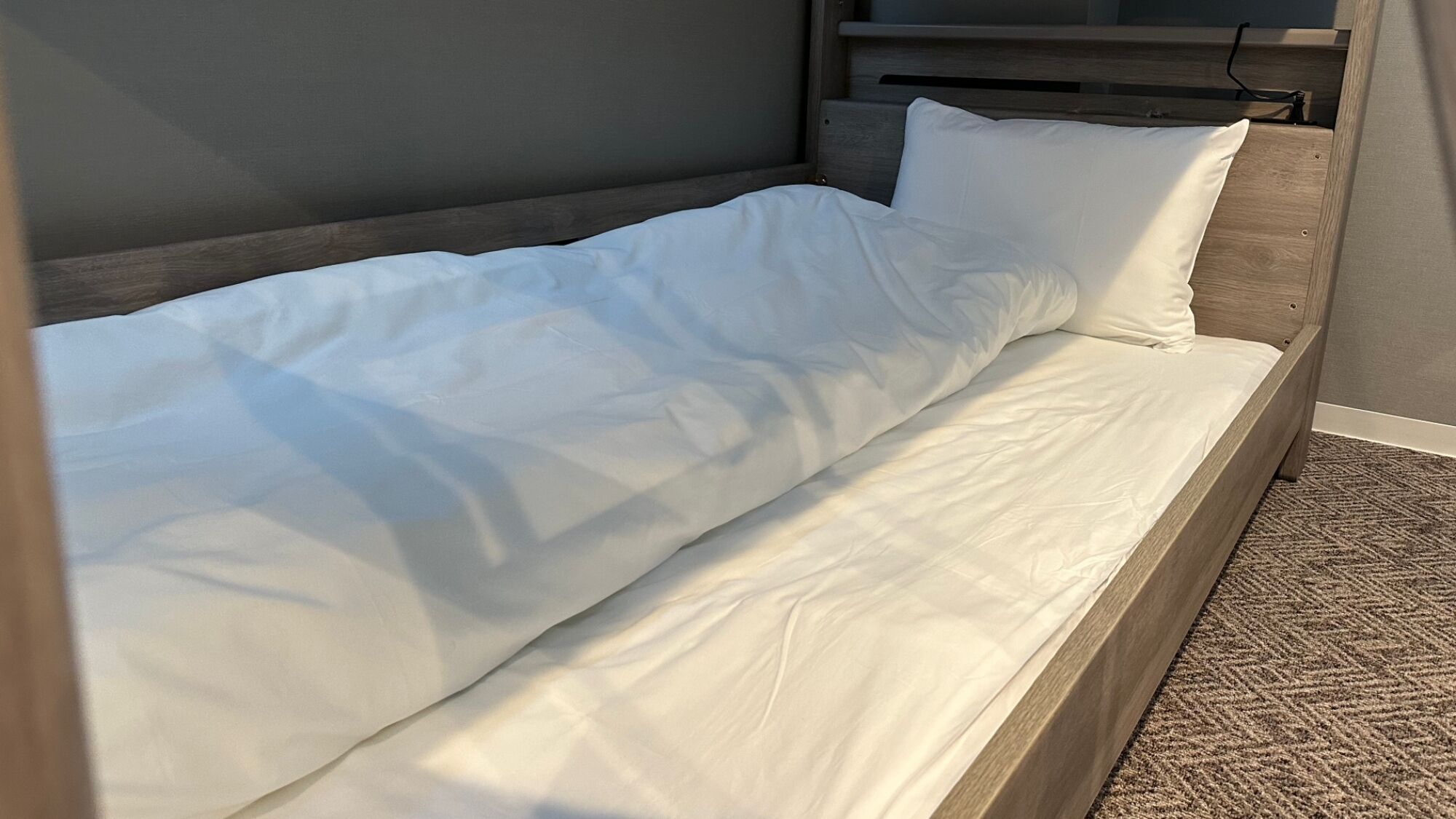 Un lit simple avec une literie blanche, dans une chambre dotée d’une tête de lit en bois et d’un tapis à motifs.