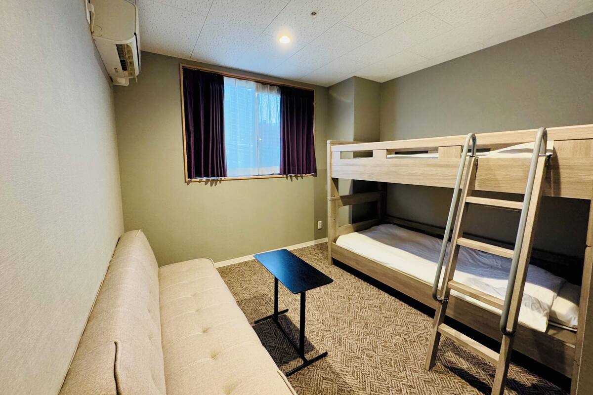A small, furnished room with bunk beds, a sofa, a small table, and a window with curtains.