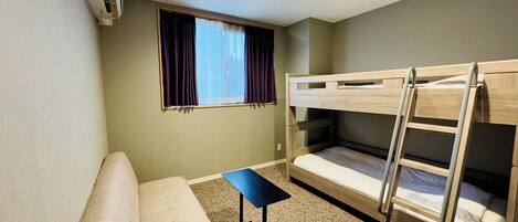 A small, furnished room with bunk beds, a sofa, a small table, and a window with curtains.