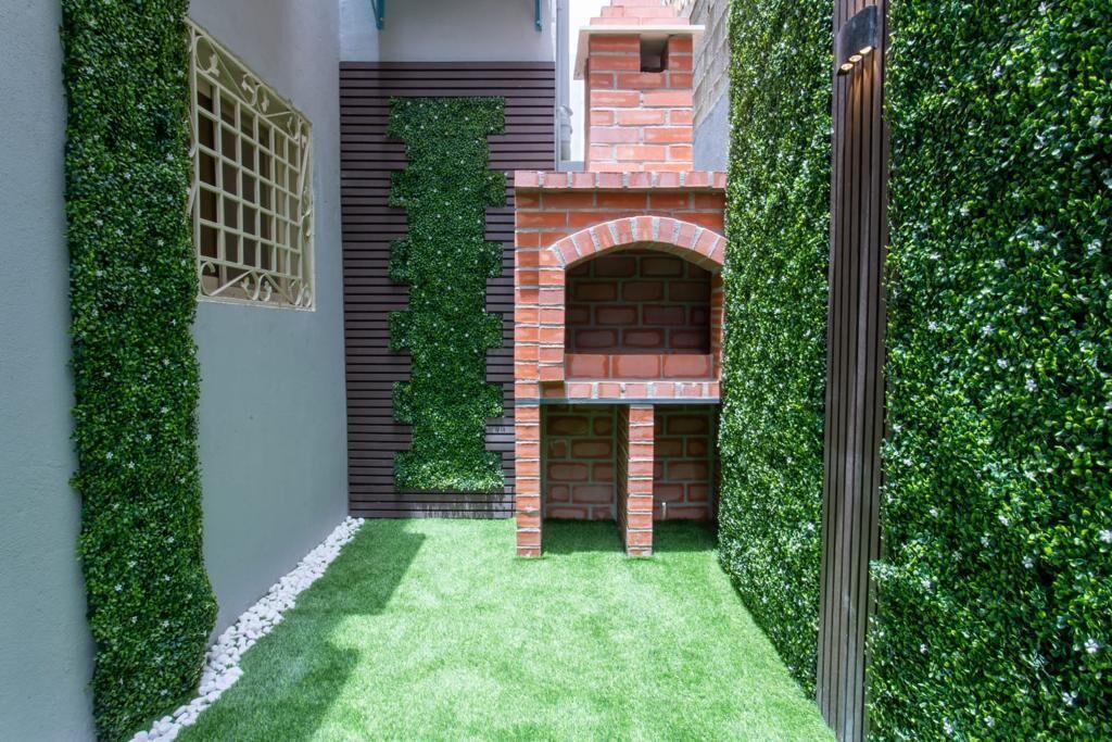Un patio estrecho con zona de barbacoa de ladrillo, rodeado de vegetación exuberante y guijarros blancos.