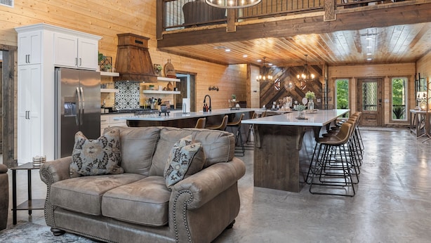 A spacious kitchen with a central island, a dining area with bar stools, a high ceiling with wooden beams, and a view of the outdoors through large windows.