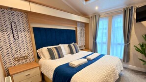 Une chambre bien rangée, avec un grand lit, une table de chevet dotée de tiroirs, un téléviseur fixé au mur et une fenêtre avec des rideaux.