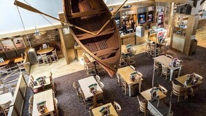 Un restaurant avec des tables et des chaises en bois, un bateau en bois suspendu au plafond et un mur de briques avec des étagères en arrière-plan.