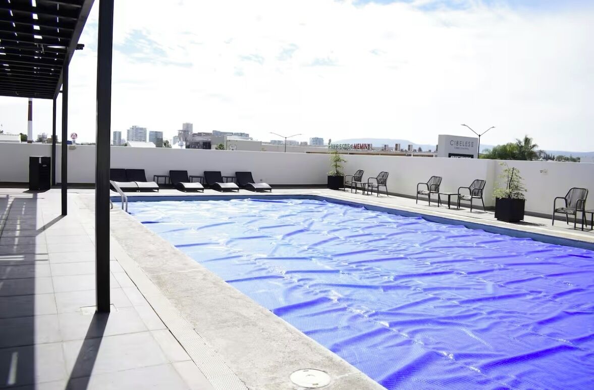 A rooftop pool with blue cover, lounge chairs, and a city view.