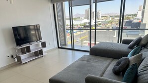 Una sala moderna con un sofá grande de color gris, un televisor de pantalla plana sobre un mueble blanco, y un ventanal que ofrece vistas a la ciudad.