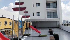 Un patio de juegos en la azotea con toboganes y estructuras para escalar, rodeado por un edificio moderno con amplias ventanas.
