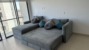 Un sofá seccional gris con varios cojines en una habitación moderna con ventanas amplias y suelo de baldosas.