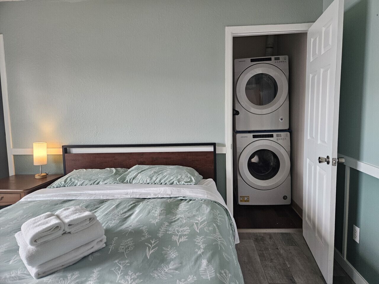 Una habitación de hotel con una cama, una lavadora y una mesita de noche con una lámpara.