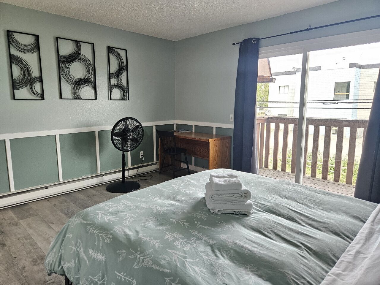 A bedroom with a bed, a desk, a chair, and a fan. There are three wall art pieces above the bed.