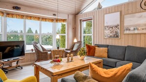 A cozy living room with a wooden ceiling, large windows, a grey sofa, a yellow armchair, and a wooden coffee table.
