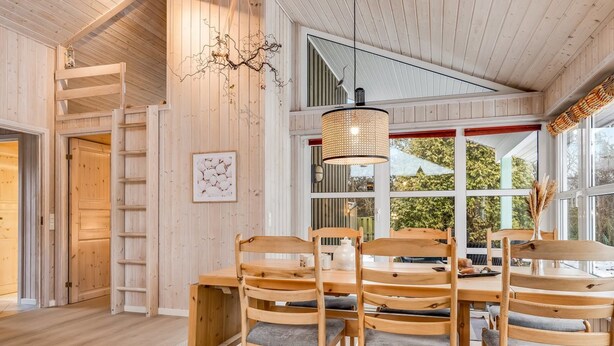 A wooden dining area with a large table and chairs, a hanging light fixture, and a view of the outdoors through large windows.