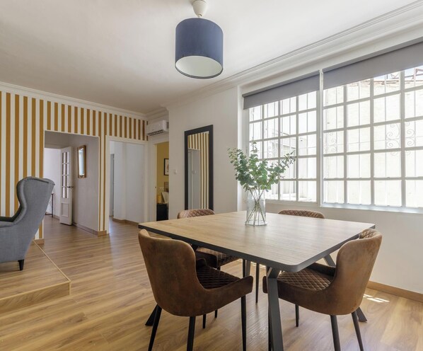 Un comedor con una mesa de madera y cuatro sillas, un jarrón con plantas verdes y un ventanal con marcos blancos.