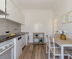Cocina compacta con lavamanos, estufa y lavadora. Hay una pequeña mesa con sillas y una planta en maceta.