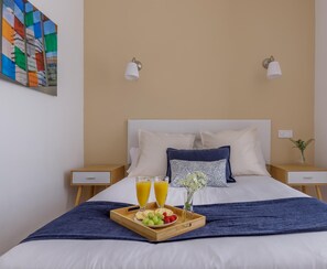 Una cama bien hecha con una manta azul, dos mesitas de noche con lámparas, una bandeja con fruta y jugo, y un cuadro abstracto de colores en la pared.