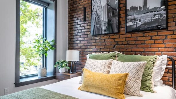 A modern bedroom with a large bed, a brick wall, and two black-and-white cityscape photographs.