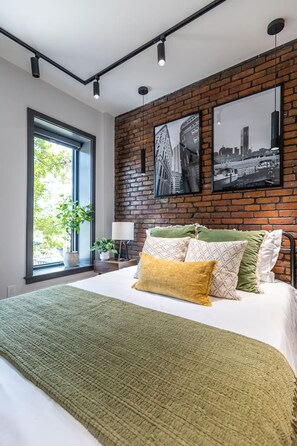 A modern bedroom with a large bed, a brick wall, and two black-and-white cityscape photographs.