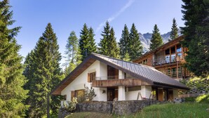 Un chalet en bois à deux étages, doté d’un toit en pente, de fondations en pierre et d’un balcon donnant sur une zone boisée.
