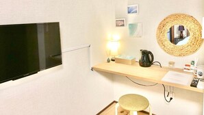 A hotel room with a flat-screen TV, a wooden shelf with a lamp, a wicker mirror, and a small stool. - ALOHADAYS Nishijin Guesthouse (Kyoto)