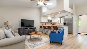 Una sala de estar moderna con un sofá, una mesa de centro y un sillón azul. Hay un televisor sobre un mueble bajo, y una zona de cocina con un refrigerador, microondas y taburetes de bar.