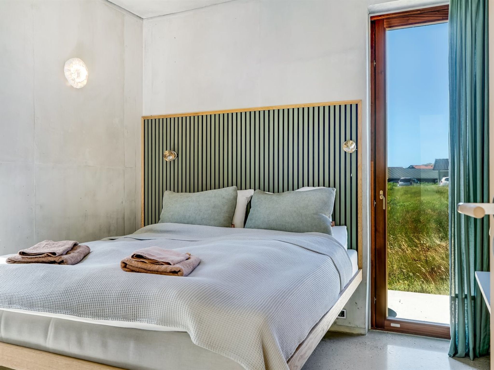 Une chambre à coucher avec un lit, des oreillers, une tête de lit et une vue sur l’extérieur, visible à travers une porte.
