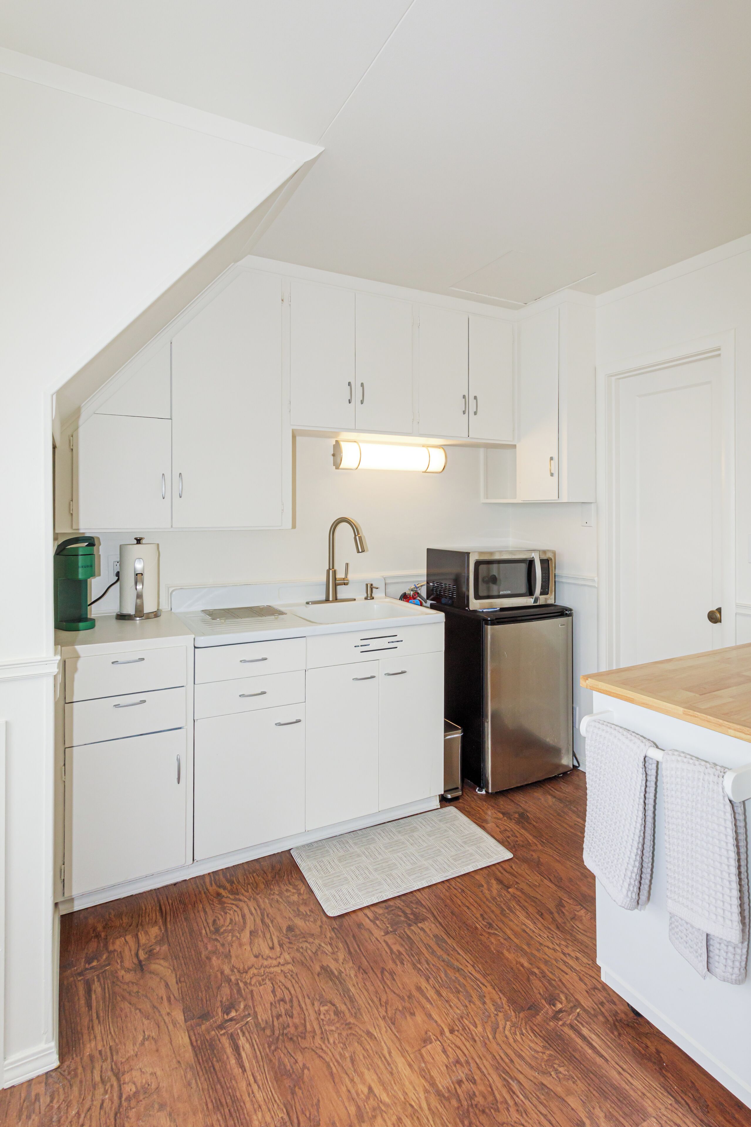 Una cocina con armarios blancos, un refrigerador de acero inoxidable, un microondas, una cafetera y una encimera de madera.