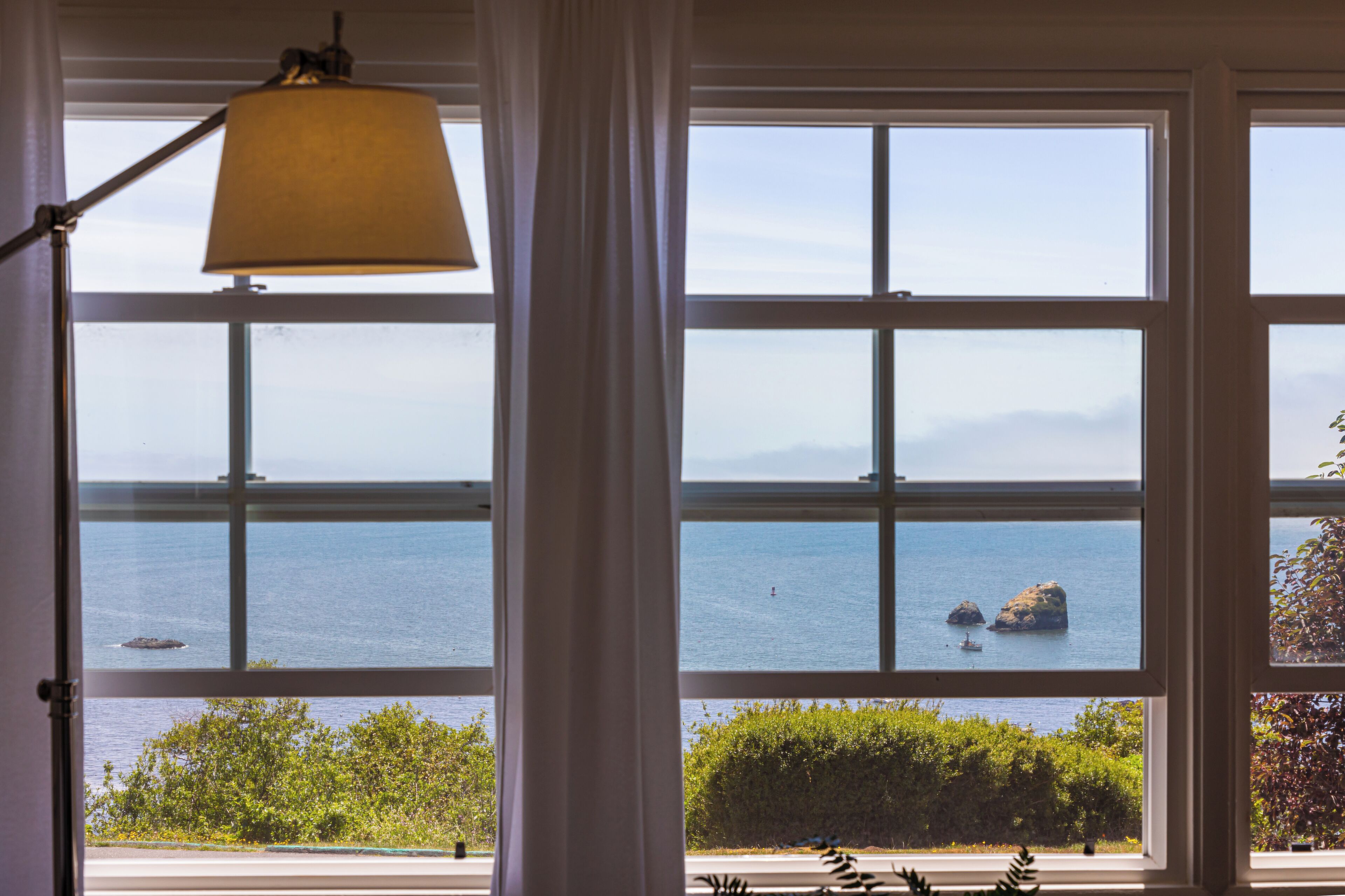 A view through a window with a lamp, a clear sky, and the sea with rocks.