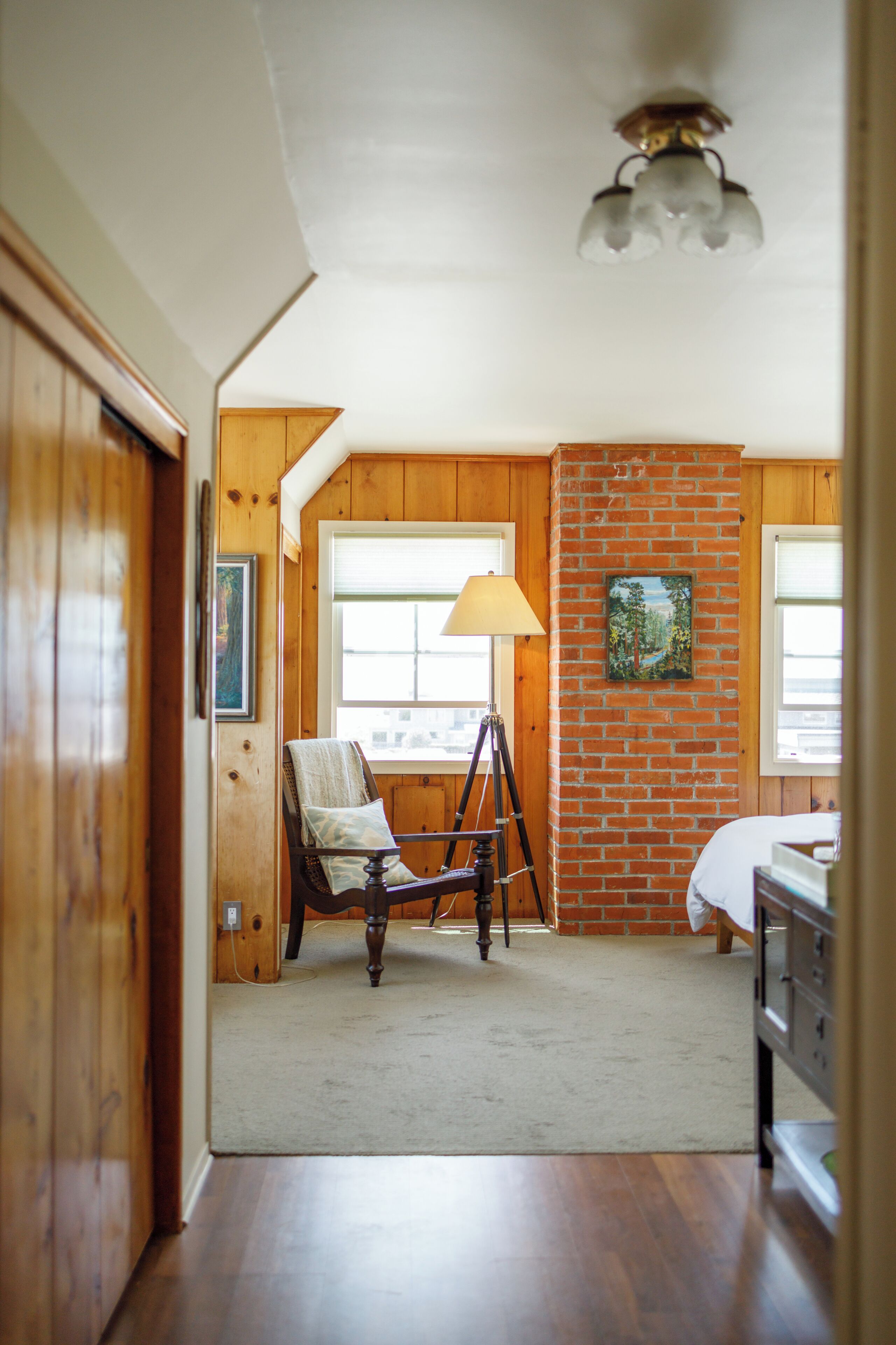 A room with wooden walls and a brick accent wall, a tripod lamp, a chair, and a bed.