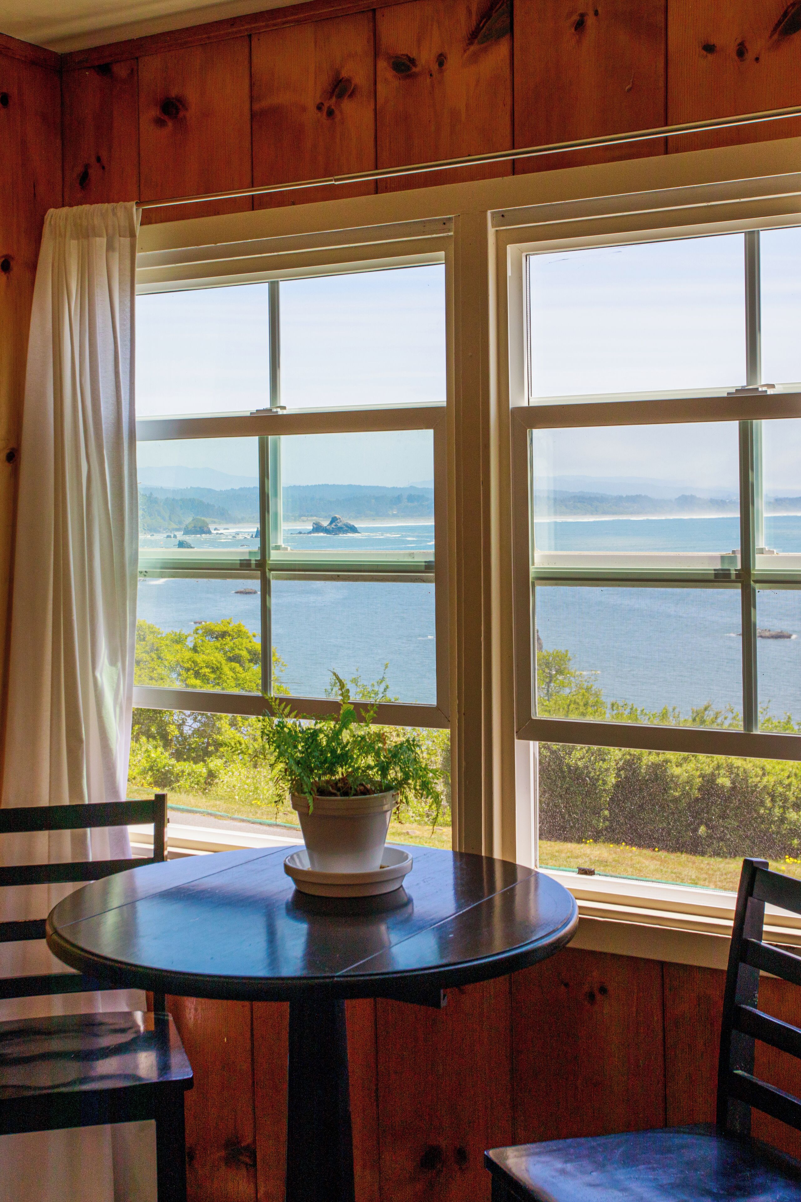 A room with a wooden interior, a round table with a potted plant, two chairs, and a view of the sea through large windows.