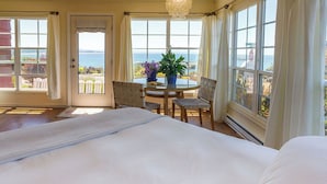 A bedroom with a large bed, a dining table with chairs, and a view of the sea through large windows.