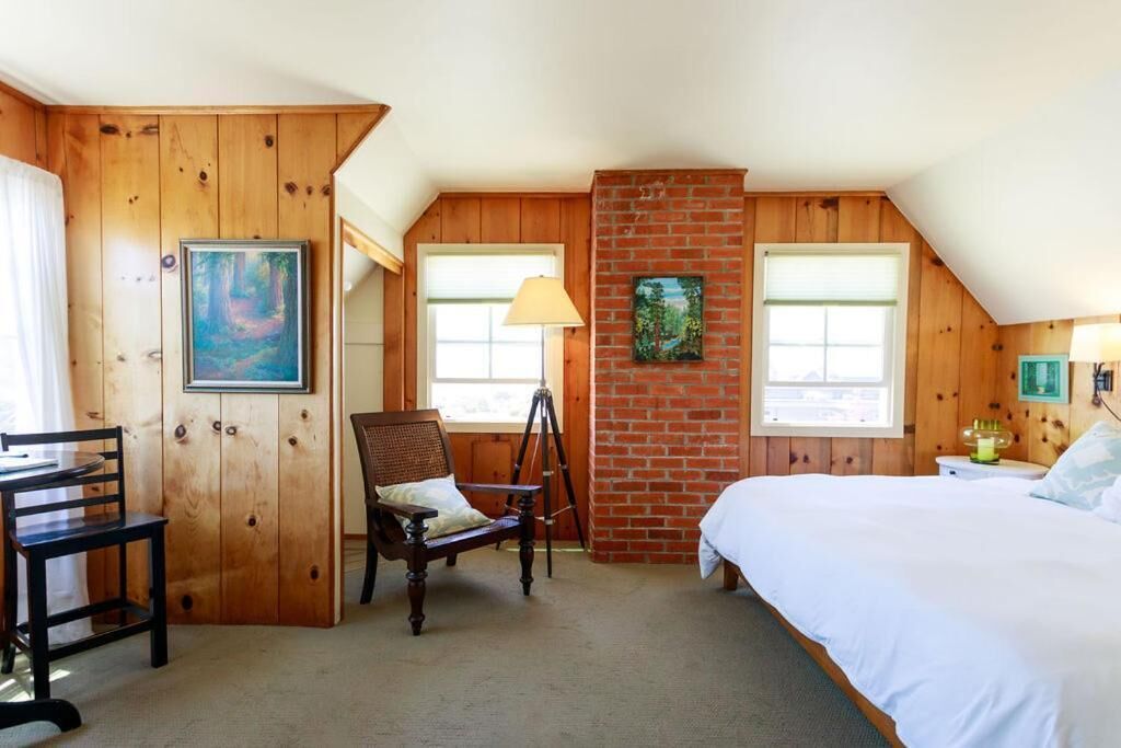 A room with a wooden paneled wall, a bed, a desk with a chair, and a tripod lamp.