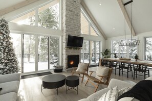 A spacious living room with a fireplace, large windows, and a high ceiling.