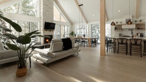 A spacious living area with a fireplace, large windows, and a dining area with a kitchen in the background. - Spa • Sauna  • Salle de jeux • Terrasse • Foyer (Les Laurentides Regional County Municipality)