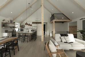 A spacious living area with a high ceiling, a dining area with a long table and chairs, and a kitchen with modern appliances and pendant lighting.
