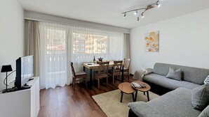 A modern living room with a grey sofa, a dining table with chairs, a TV, and a painting on the wall.