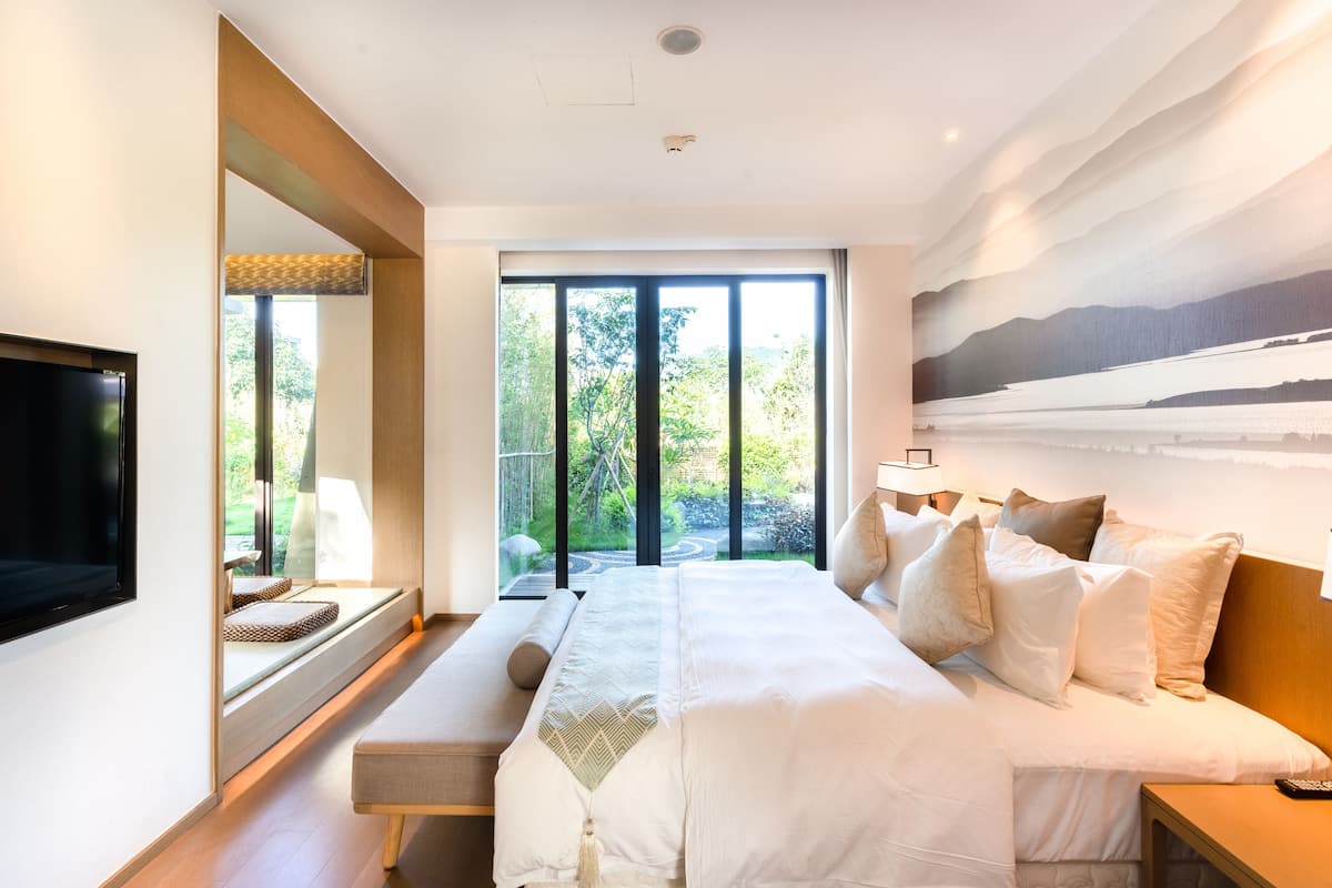 A modern bedroom with a large bed, a TV, and a view of greenery through large windows.