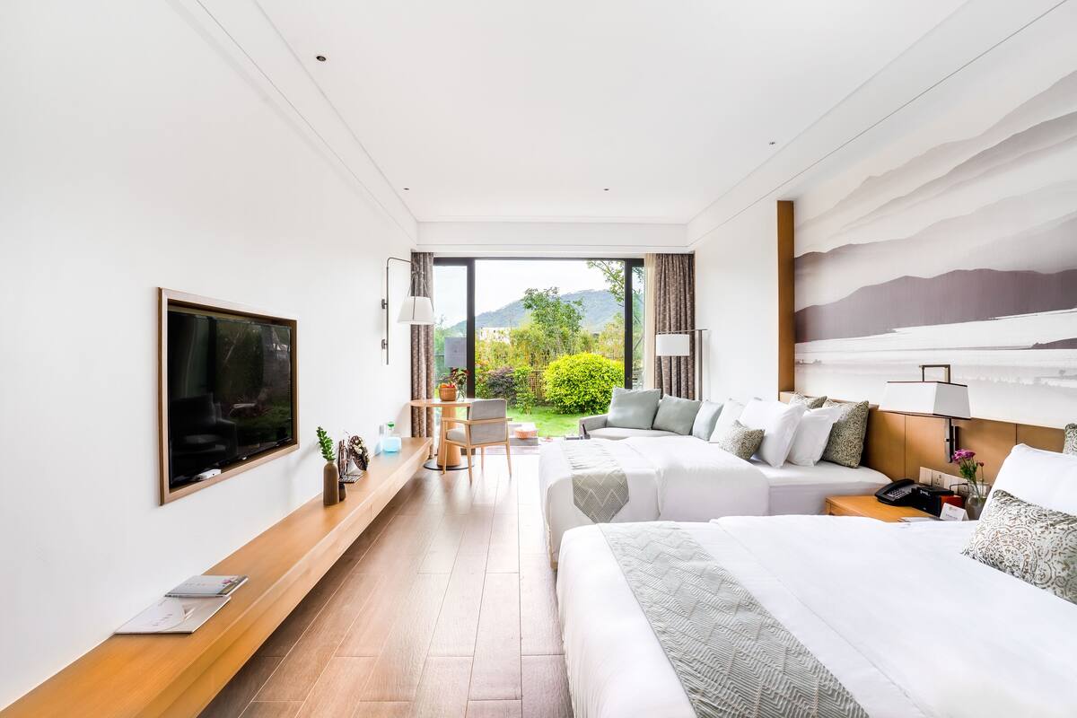 A modern hotel room with a large bed, a desk, and a view of greenery and mountains.