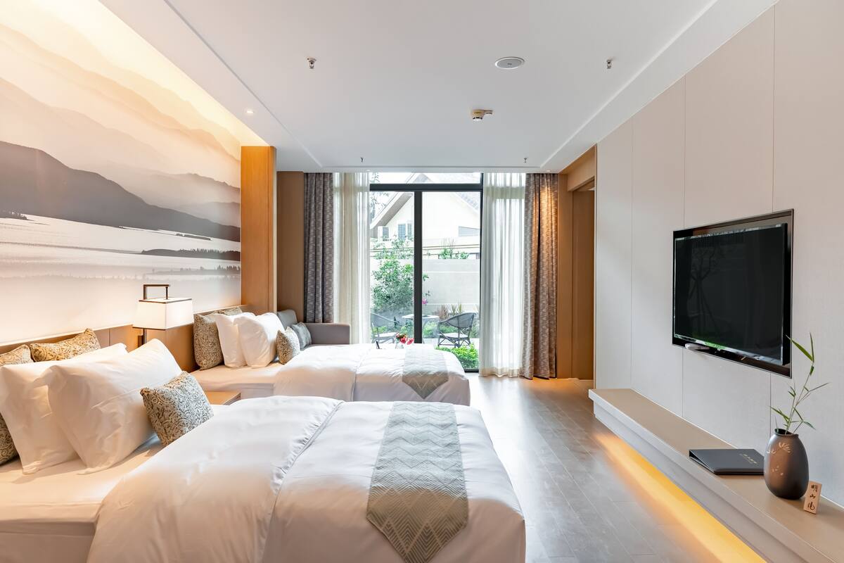 A modern hotel room with two beds, a large window, a flat-screen TV, and a bedside table with a vase.