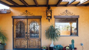 Une porte à deux battants en bois, ornée de motifs métalliques complexes, un pot de fleurs et divers cactus devant un bâtiment dont la façade est recouverte d’un enduit jaune et qui possède une fenêtre.