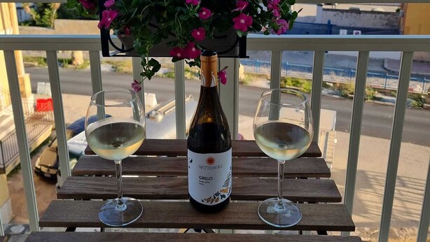A balcony with a wooden table, a bottle of wine, two wine glasses, and a potted plant with pink flowers.