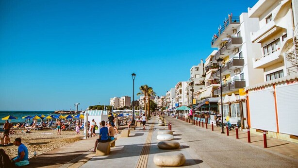 A coastal promenade with beach umbrellas, people, and buildings along the seaside. - Lazareta Studio | Nea Chora (Chania)