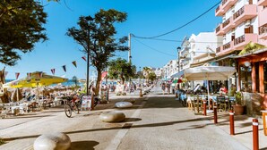 A sunny street lined with outdoor cafes, tables, and chairs, with buildings on either side and a clear blue sky. - Lazareta Studio | Nea Chora (Chania)