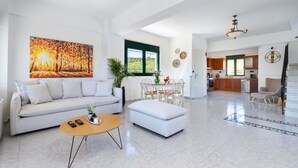 A spacious living area with a sofa, coffee table, and dining table. A kitchen is visible in the background. - Villa Giannari | Stalos (Chania)