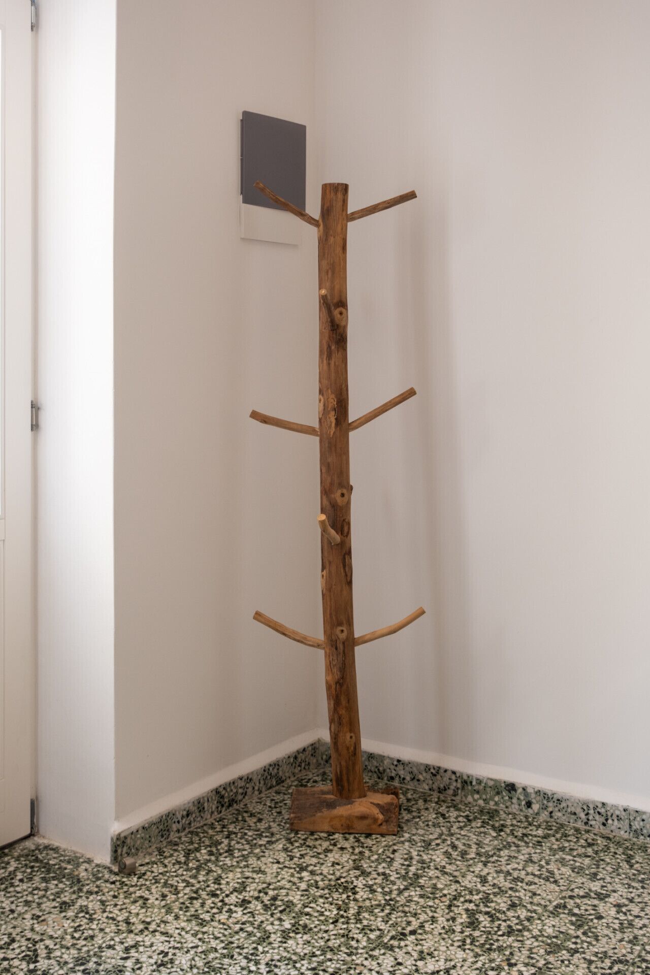 A wooden coat rack in a corner with a granite floor and a white door.