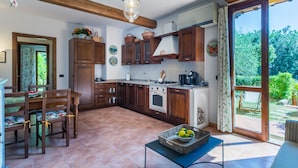 A kitchen with wooden cabinets, a dining table with chairs, a blue table with a fruit basket, and a view of the garden through the glass door. - Il Castello by Interhome (Montescudaio)