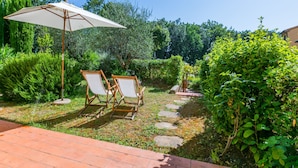 A garden patio with two wooden chairs under a white umbrella, surrounded by lush greenery and a stone pathway. - Il Castello by Interhome (Montescudaio)